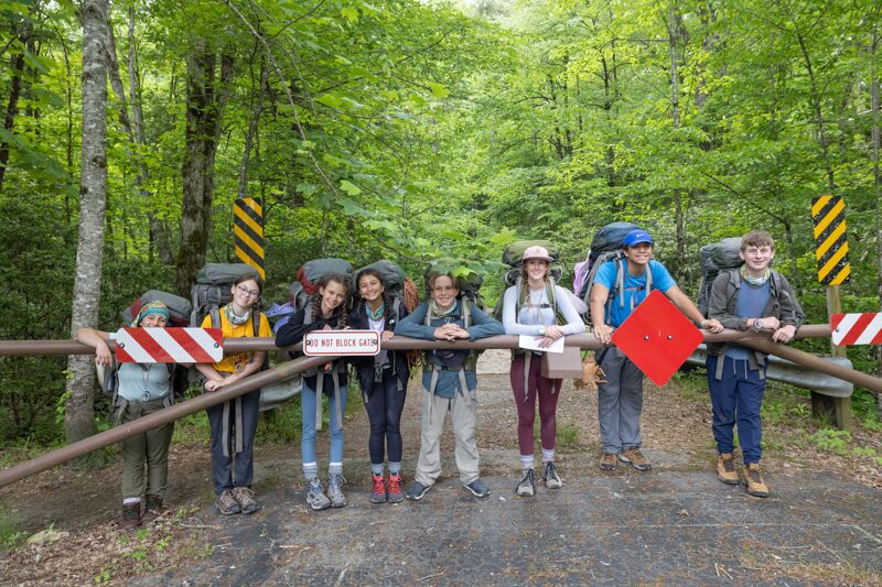 The image shows a group of seven young people standing behind a metal barrier in a wooded area. They are all wearing backpacks, suggesting they are hikers or campers. The group is diverse in appearance, and they are all looking towards the camera. The background is filled with lush green trees, indicating a natural setting. The overall impression is one of a group of friends enjoying an outdoor adventure.